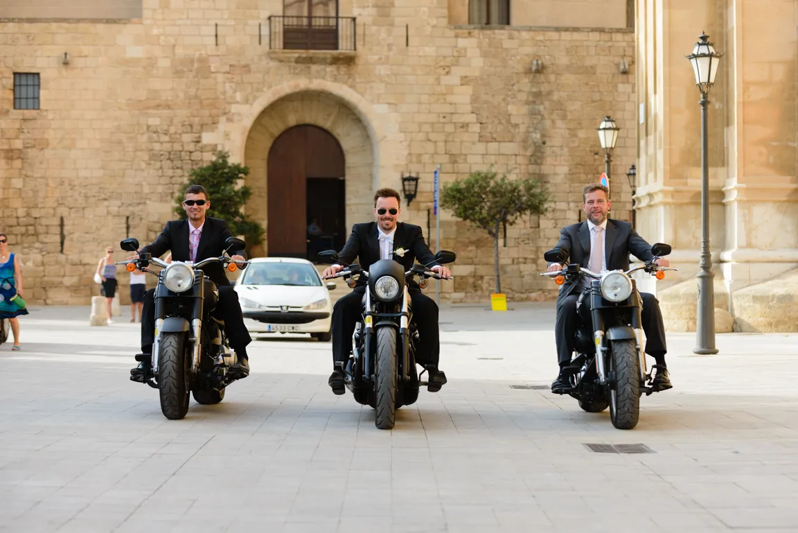 Three men in suits riding black motorcycles on a paved street with a stone building in the background.