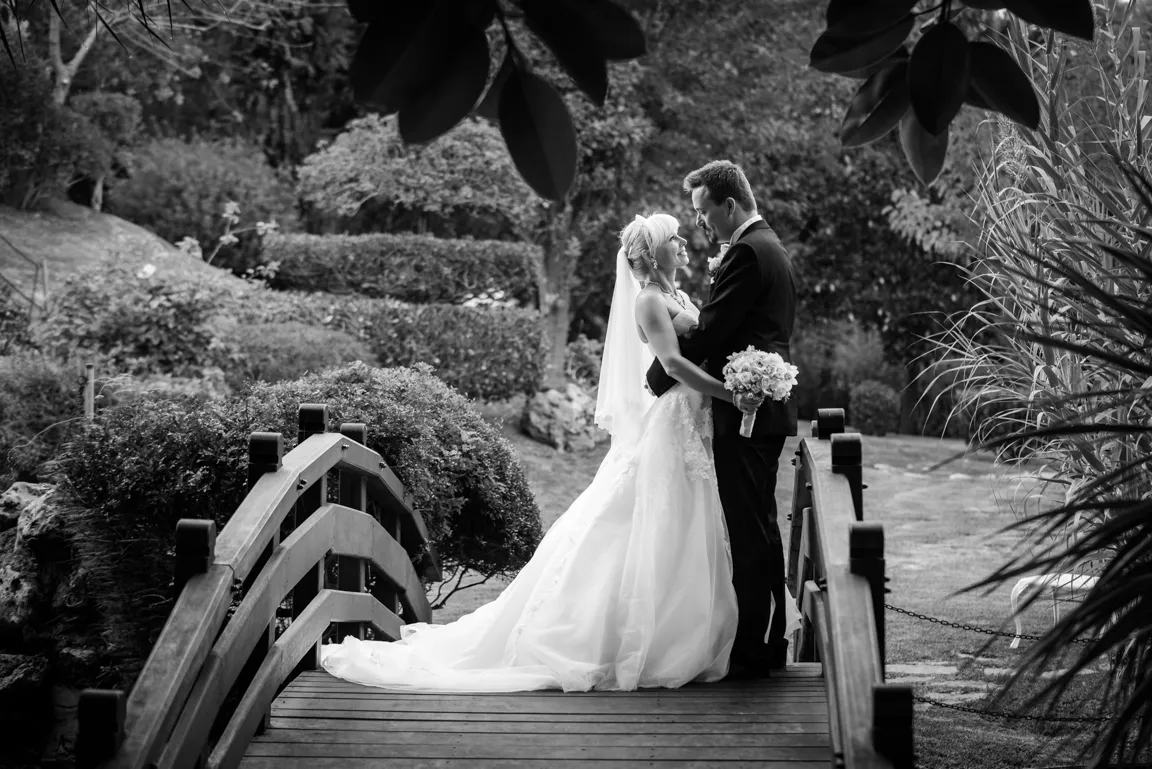 Bride and groom embracing on a wooden garden bridge surrounded by lush greenery in a black and white wedding photograph.