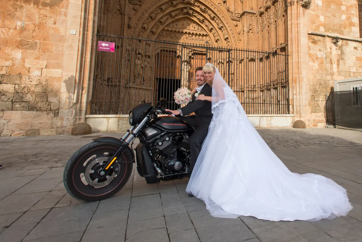 Bride in white wedding dress and groom in suit posing on a black Harley-Davidson motorcycle in front of an ornate stone building entrance.