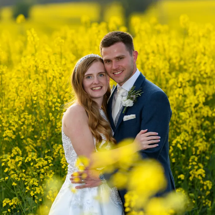 Smiling bride in a white dress and groom in a blue suit embracing in a bright yellow flower field.