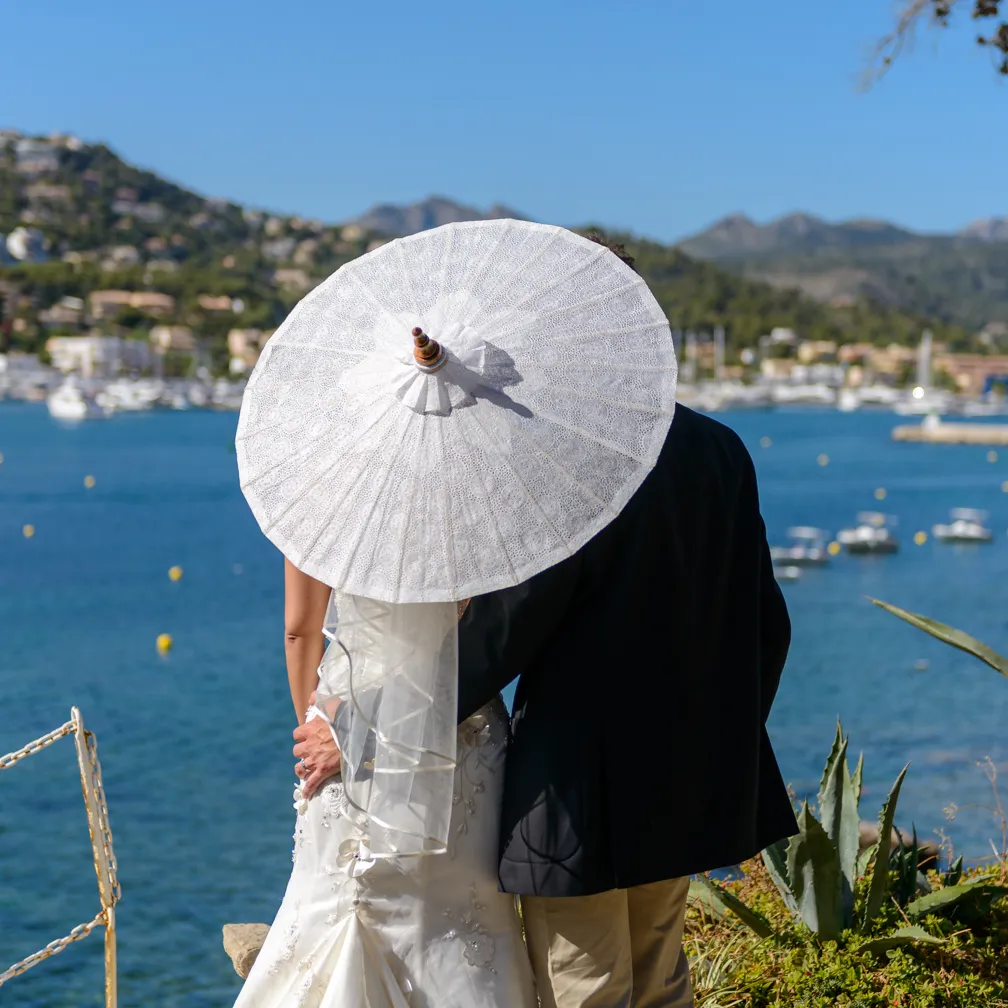 Bride and groom standing by a waterfront with bride holding a white parasol behind their backs.