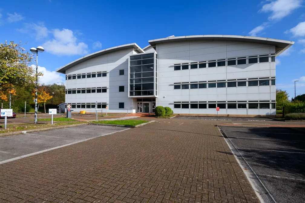 Modern three-story office building with large glass entrance and adjacent empty parking lot under a blue sky.