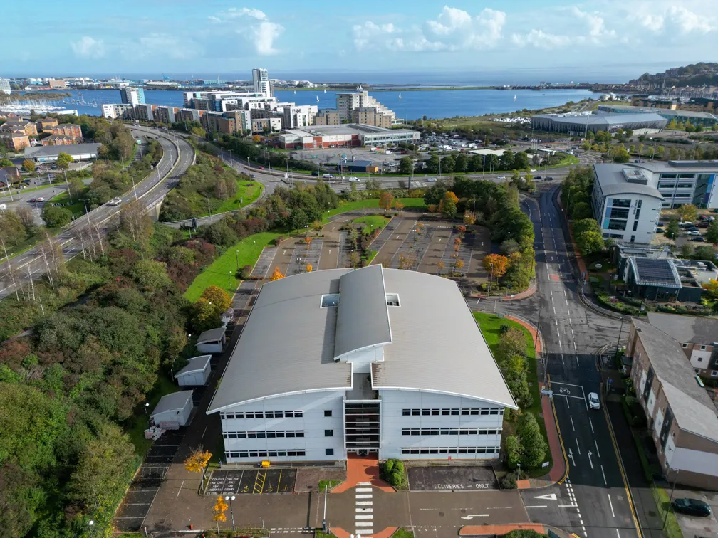 Aerial view of a modern, curved-roof office building surrounded by parking lots, roads, green trees, and a coastal cityscape in the background.