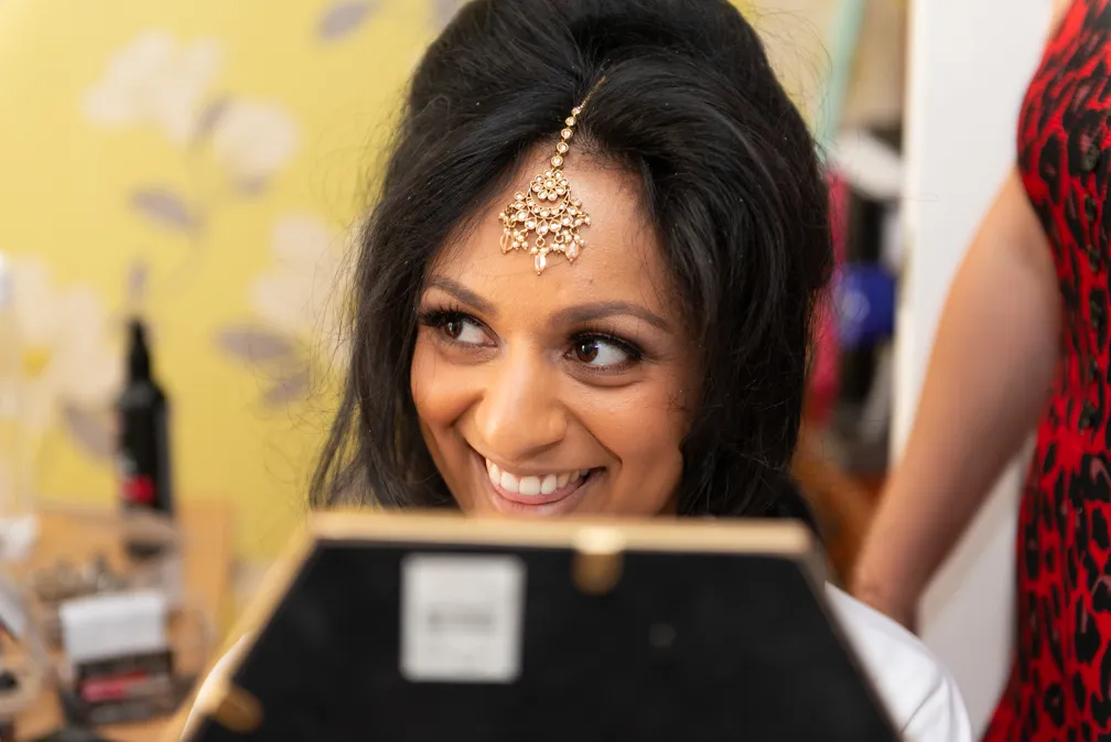 Smiling woman with dark hair wearing a traditional gold headpiece, looking into a mirror.
