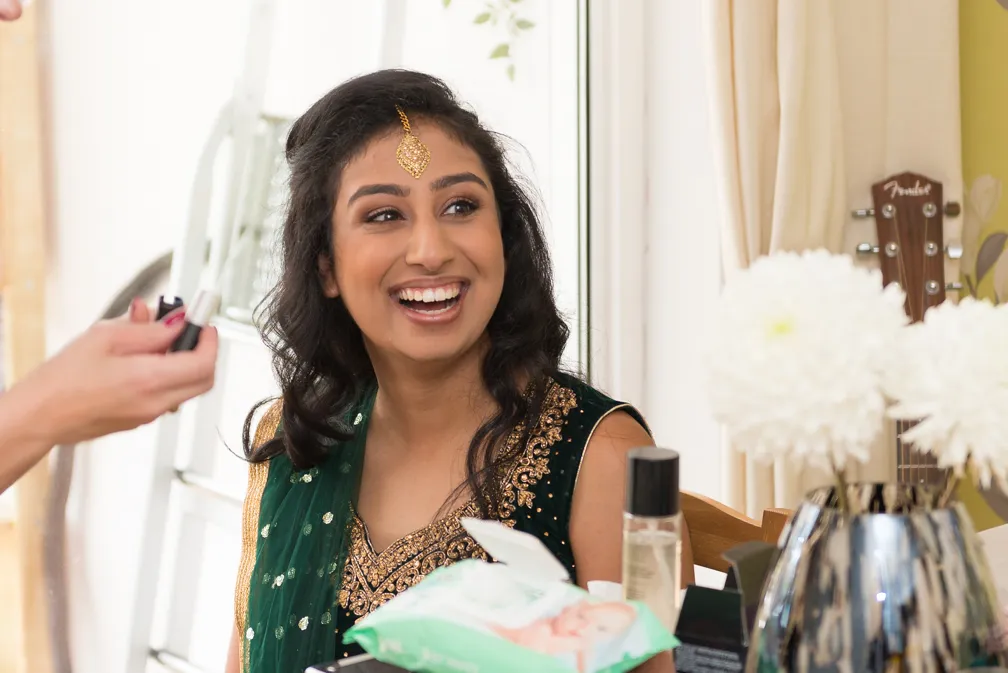 Smiling woman wearing traditional Indian attire and jewelry getting makeup applied in a bright room.