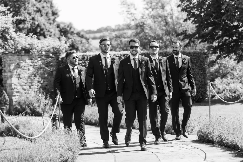 Five men in suits and sunglasses walking confidently on a paved garden path.