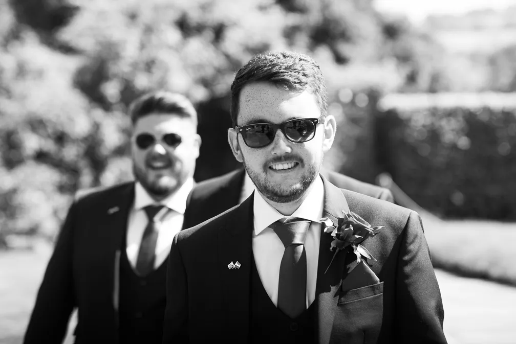 Smiling man in a suit with boutonniere and sunglasses standing outdoors with a blurred man in a suit behind him.