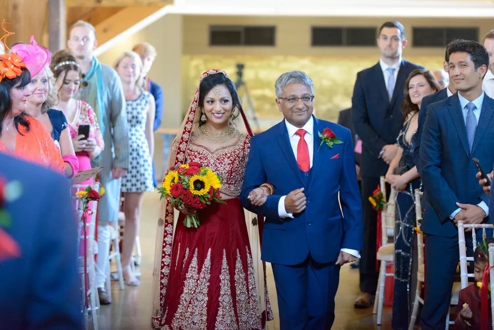 Bride in traditional red and gold attire holding a bouquet of sunflowers and red roses, walking down the aisle arm-in-arm with a man in a blue suit and red tie.