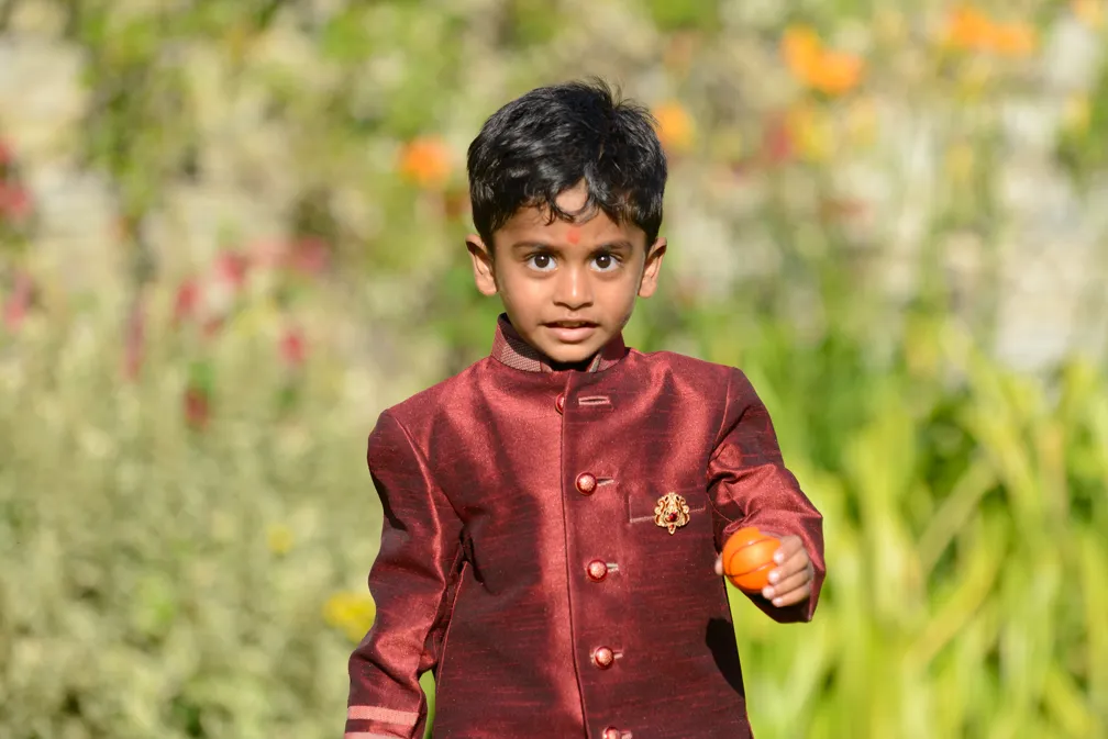 Young boy wearing a maroon traditional outfit holding a small orange ball outdoors with blurred greenery background.