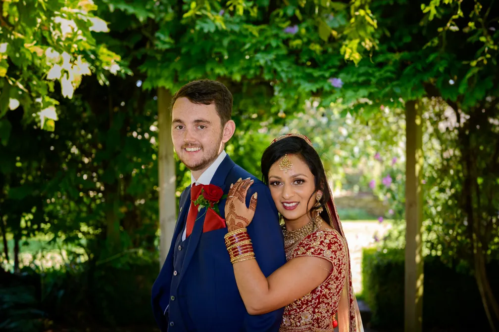 Bride in traditional red and gold attire hugging groom in blue suit with red rose boutonniere under leafy pergola.
