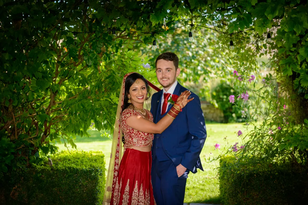 Bride in red and gold traditional attire with henna embraces groom in blue suit under green leafy arch.