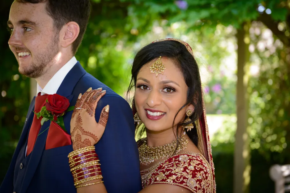 Bride in traditional red and gold Indian attire with henna on her hand, smiling and embracing groom in a blue suit with a red rose boutonniere outdoors.