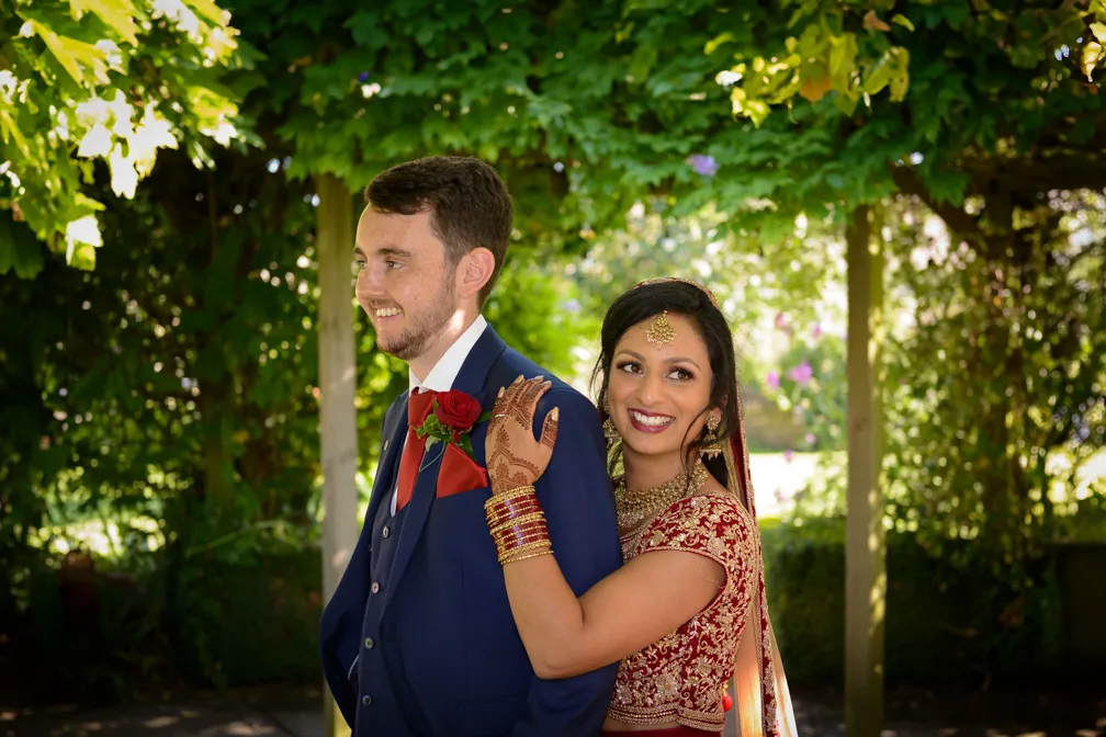 Bride in traditional red and gold attire hugging smiling groom in navy blue suit with red tie and rose boutonniere under green leafy arch.