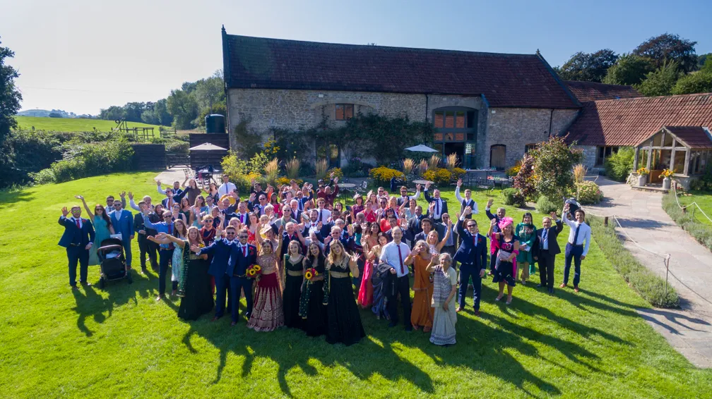 Large group of people dressed in formal attire standing on green lawn outside rustic stone building, smiling and waving at the camera on a sunny day.
