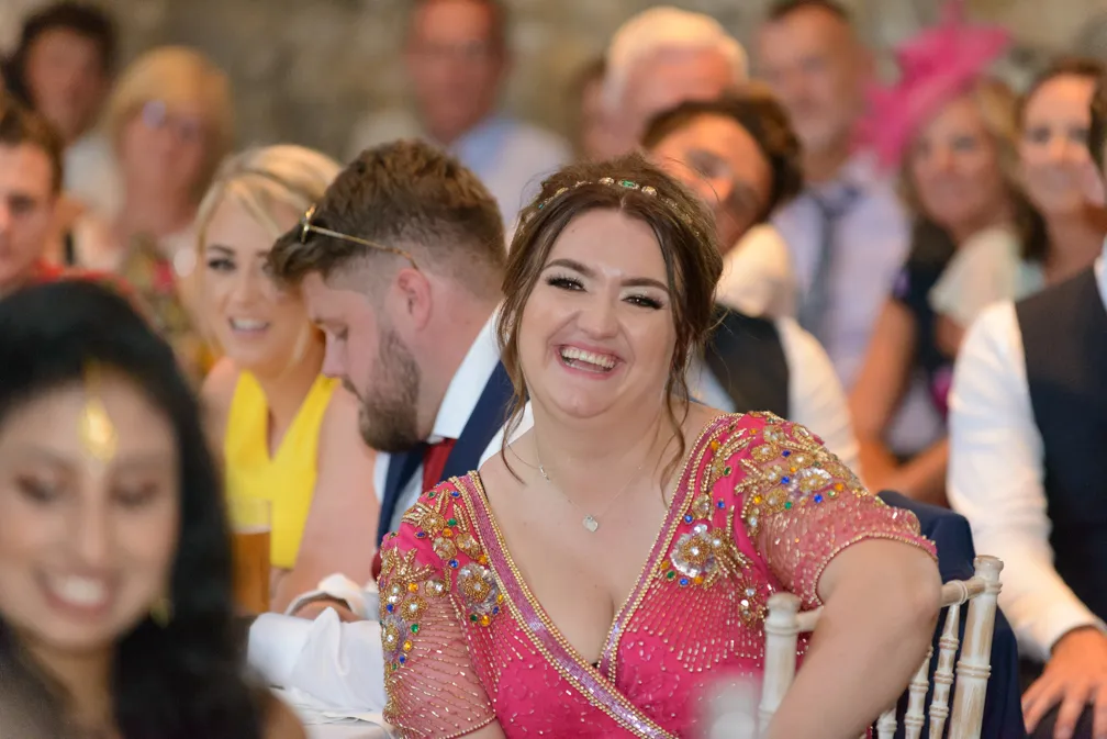 Smiling woman in a pink embellished dress laughing at an event with seated guests in the background.