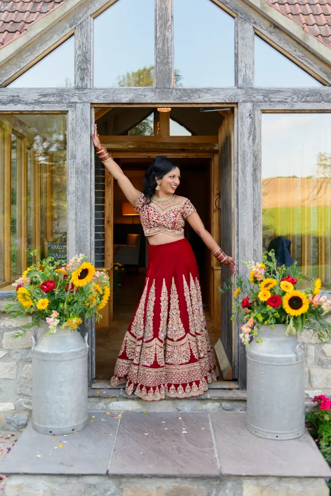 Woman in a red and gold traditional outfit posing in a wooden framed doorway flanked by vases of colorful flowers.