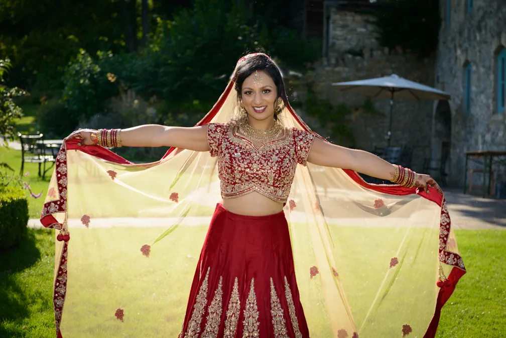 Woman in red traditional Indian attire with gold embroidery holding out sheer veiled dupatta outdoors in a garden.