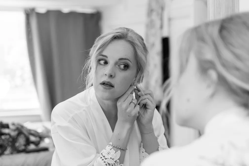 Woman wearing a robe putting on an earring while looking in a mirror.