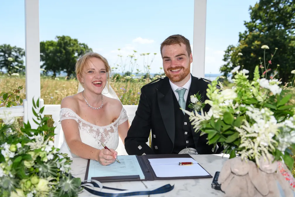Bride and groom smiling while signing wedding documents at a table with floral arrangements, outdoors on a sunny day.