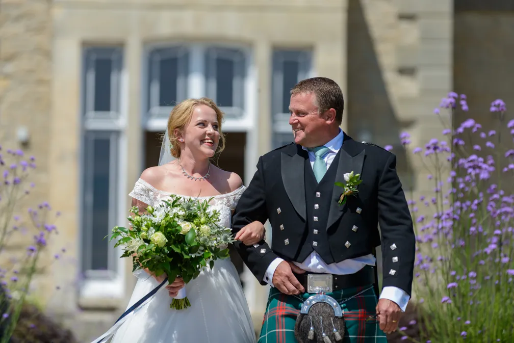 Bride and groom smiling and walking arm in arm outdoors, with the groom wearing a traditional Scottish kilt outfit.