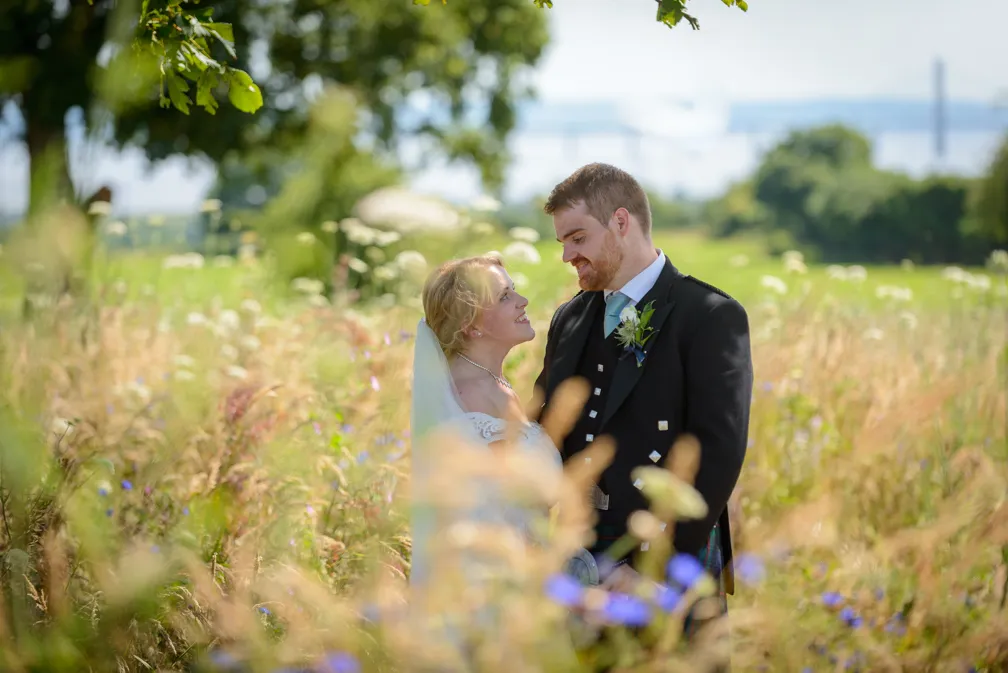 Bride and groom smiling at each other in a sunlit meadow with wildflowers and greenery.