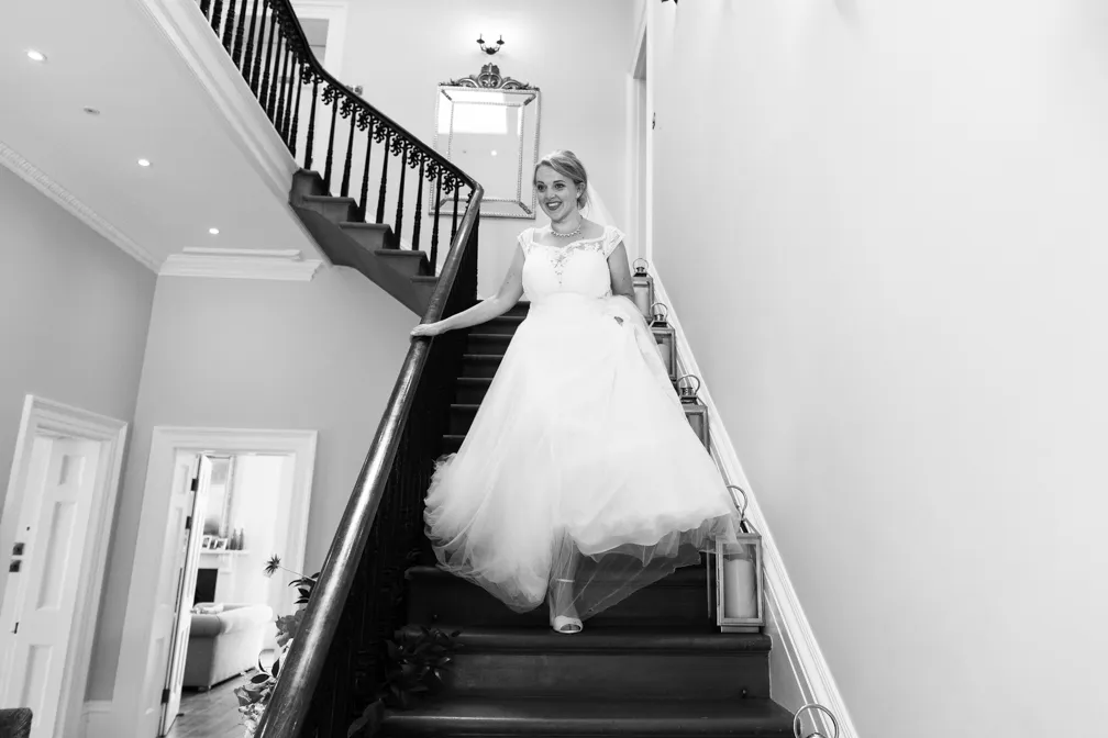 Smiling bride in a flowing wedding gown descending a staircase indoors.