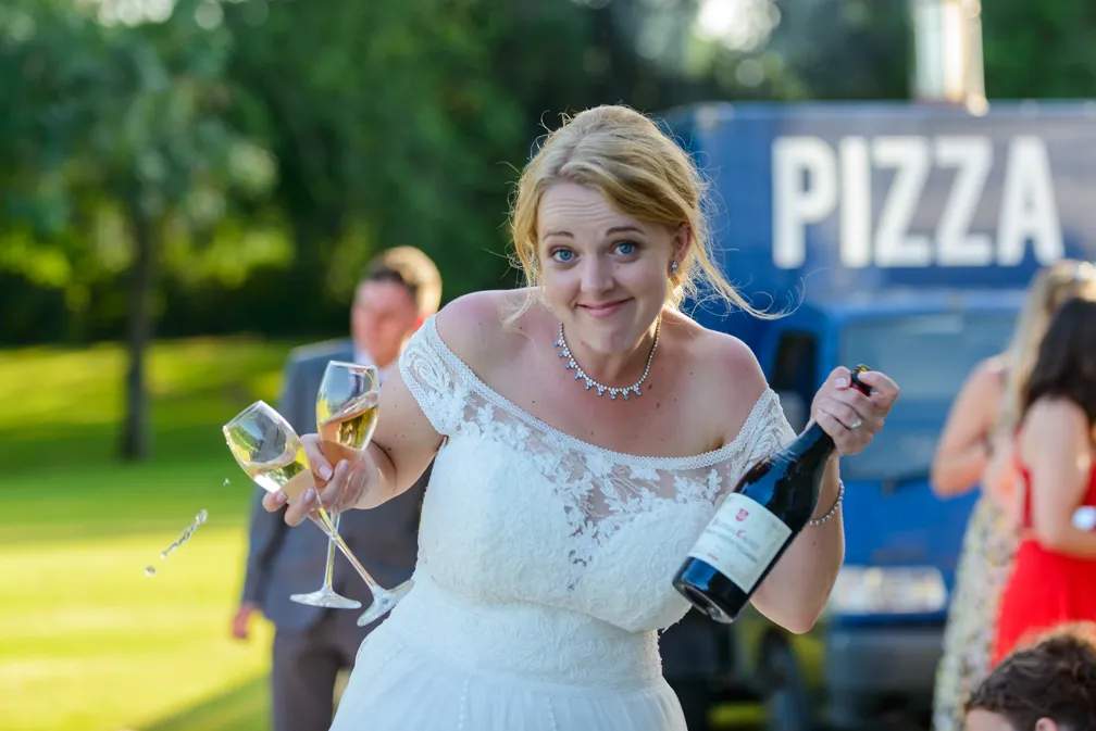Bride in white wedding dress holding two champagne glasses and a bottle with a blue truck labeled 'PIZZA' in the background.