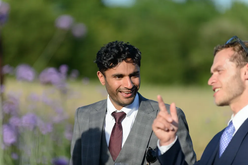 Two men in suits chatting outdoors with purple flowers and greenery in the background.