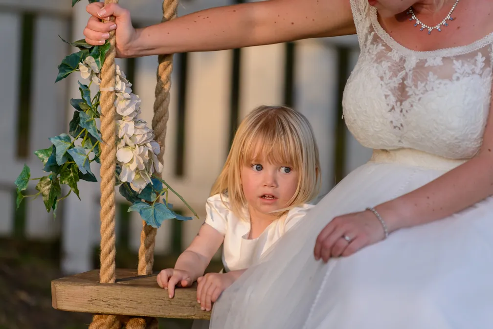 Young child in white dress sitting on a swing next to a woman in a wedding gown holding the swing ropes decorated with green leaves and white flowers.