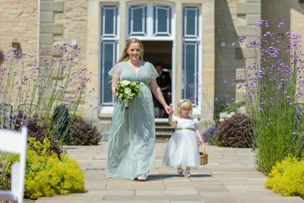 Woman in light green dress holding bouquet and hand of a young girl in a white dress walking down a stone path lined with purple flowers outdoors.