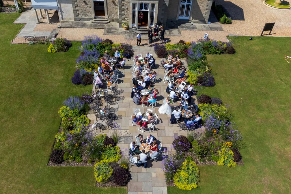 Aerial view of guests seated at tables on a garden patio outside a stone building, with vibrant flower beds surrounding the area.