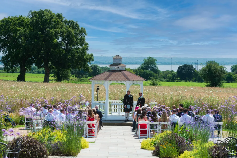 Outdoor wedding ceremony in front of a white gazebo with two men in kilts and audience seated on white chairs in a garden with fields and large trees in the background.