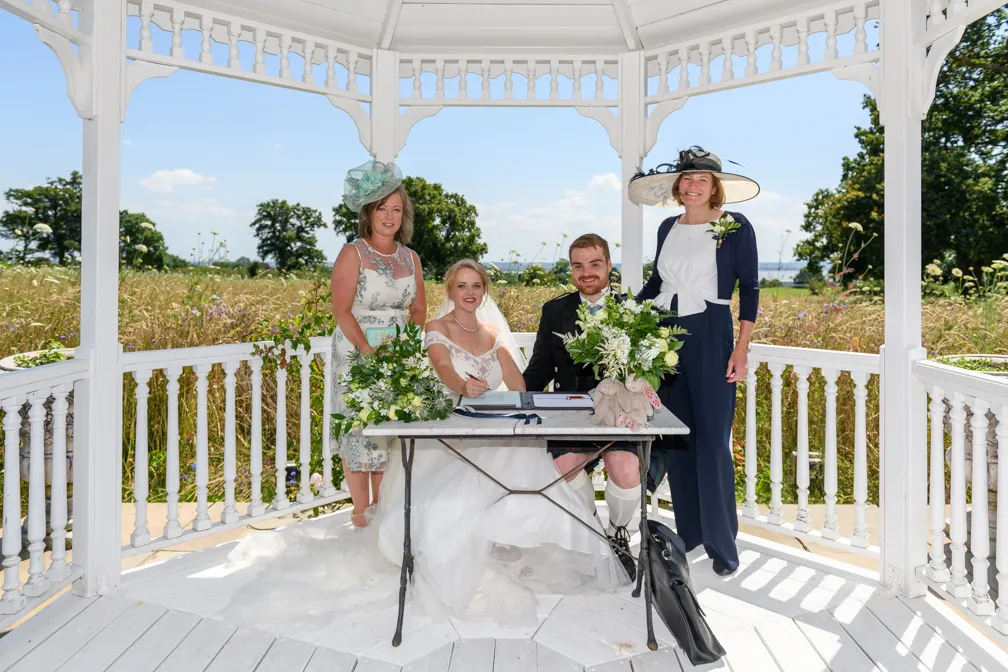 Bride and groom sitting at a table signing a document under a white gazebo with two women standing behind them, one in a blue and white dress and wide-brimmed hat and the other in a floral dress with a teal fascinator.