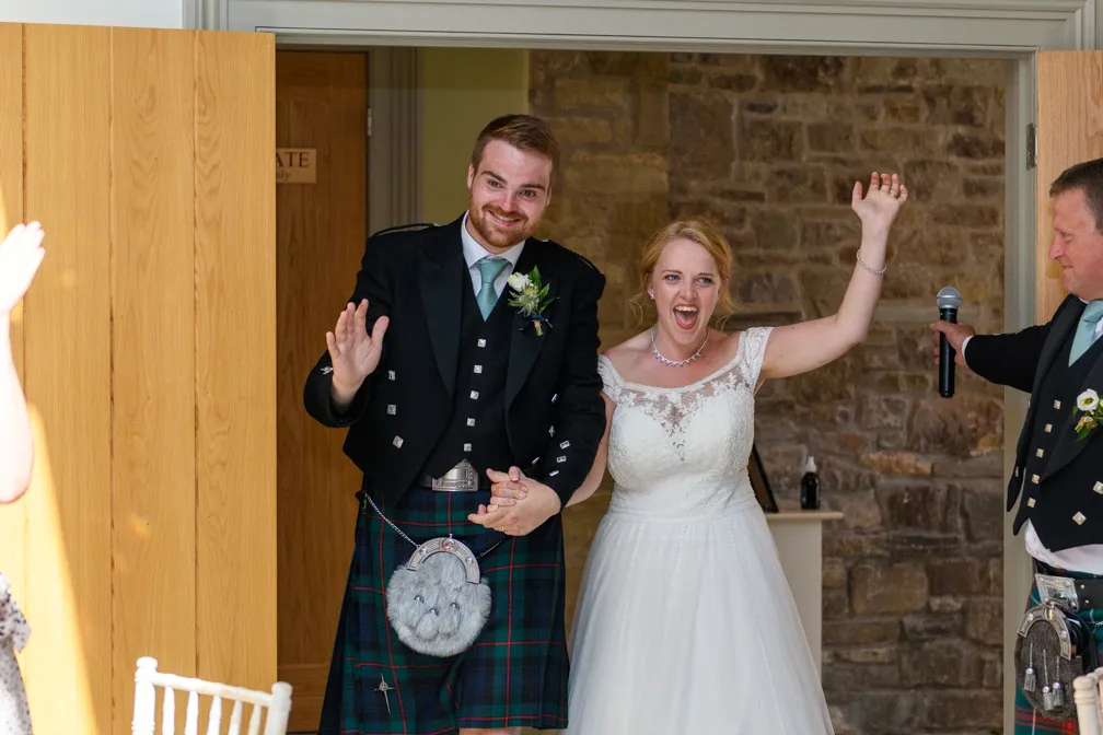 Bride and groom joyfully entering a room, holding hands; groom is wearing a traditional Scottish kilt and bride is in a white lace wedding dress.