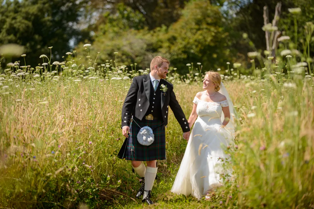Bride in white off-shoulder gown and groom in traditional Scottish kilt walking hand in hand through a sunlit meadow with tall grass and wildflowers.