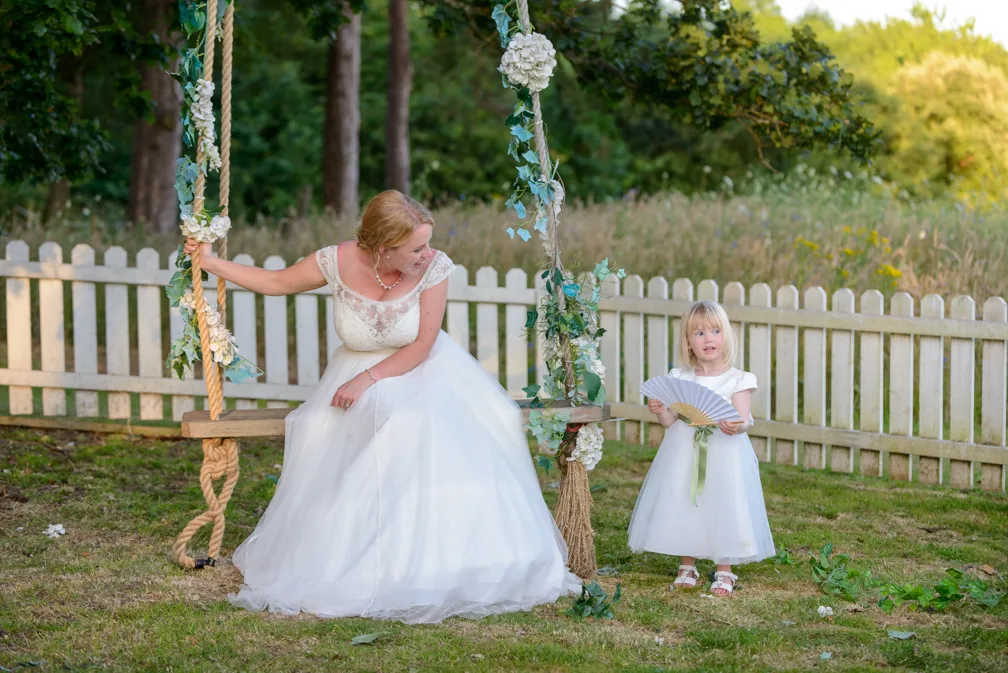 Bride in white dress sitting on a flower-adorned wooden swing, smiling at a young girl in a white dress holding a folding fan in a garden with a wooden fence.