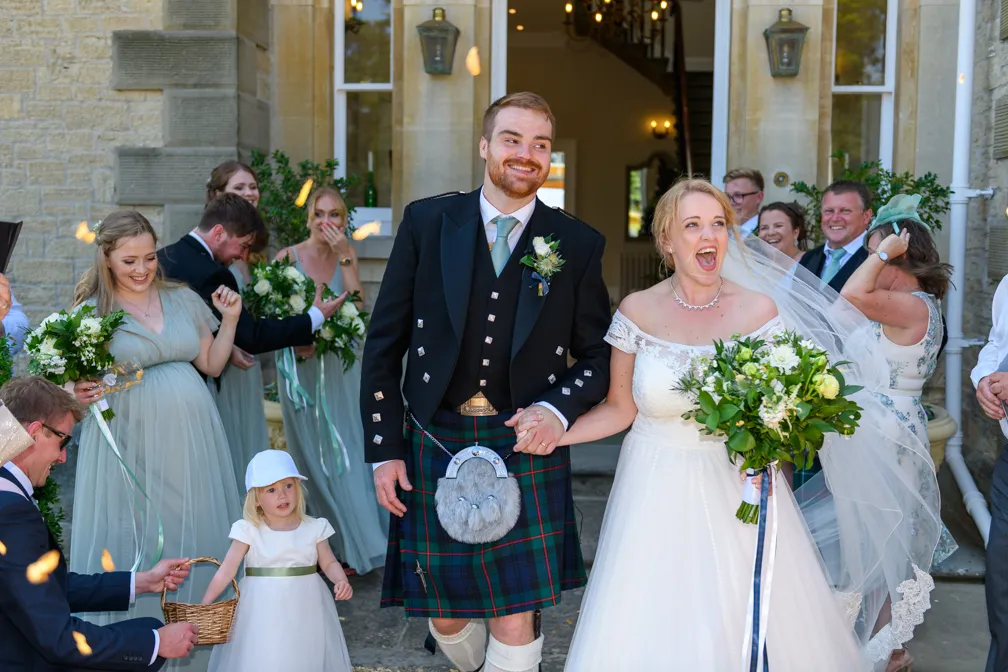 Bride and groom holding hands and smiling, surrounded by bridesmaids and guests throwing confetti outside a stone building.