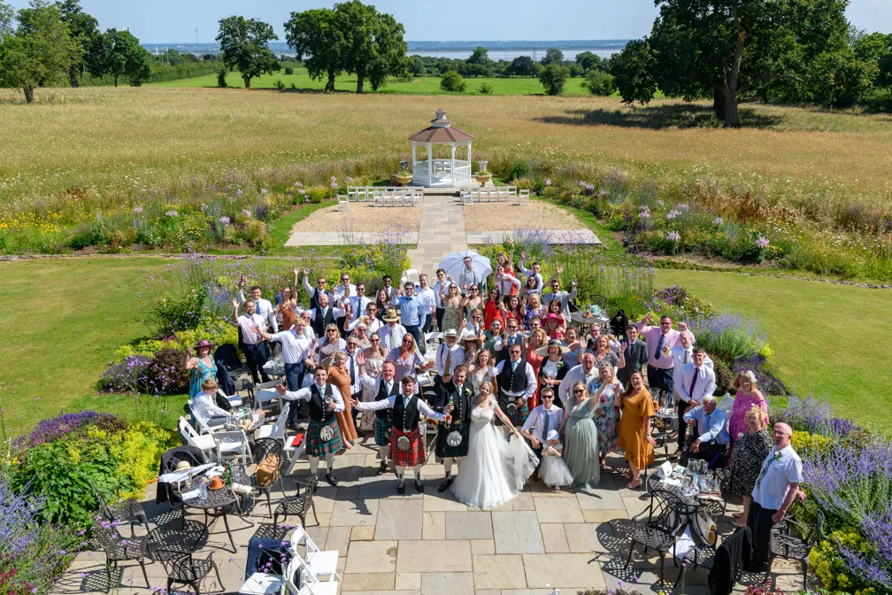 Large outdoor wedding group photo with bride, groom, and guests gathered on a stone patio in front of a small white gazebo surrounded by greenery and flowers.