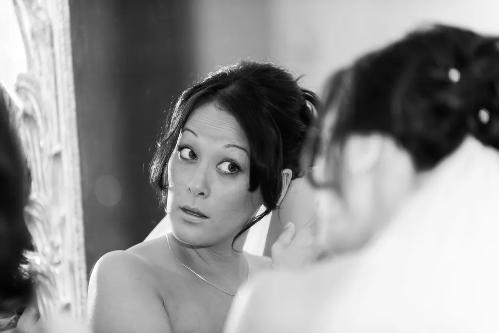 A woman with dark hair styled up looks into a mirror with a surprised expression in black and white photo.