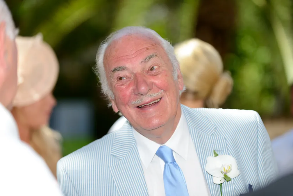 Smiling elderly man in a light blue striped suit and blue tie with a white flower boutonniere at an outdoor event.