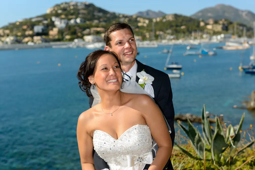 Smiling bride in a beaded white strapless wedding dress and groom in dark suit with white boutonniere posing by a coastal bay with boats and hills in the background.