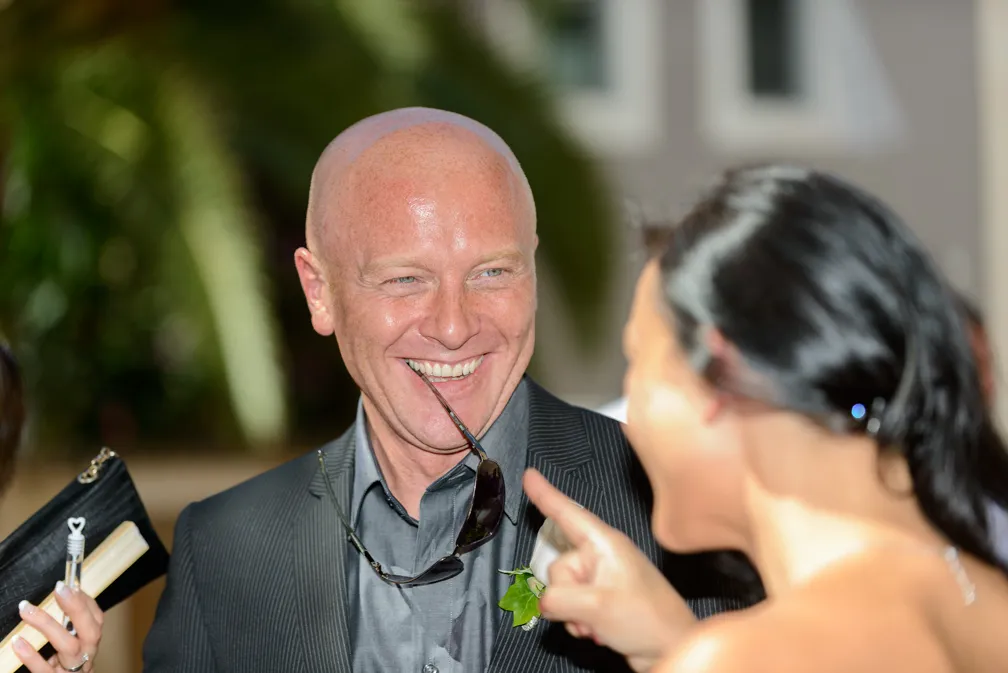 Smiling bald man in a suit holding sunglasses in his mouth, interacting with a woman pointing her finger at him.