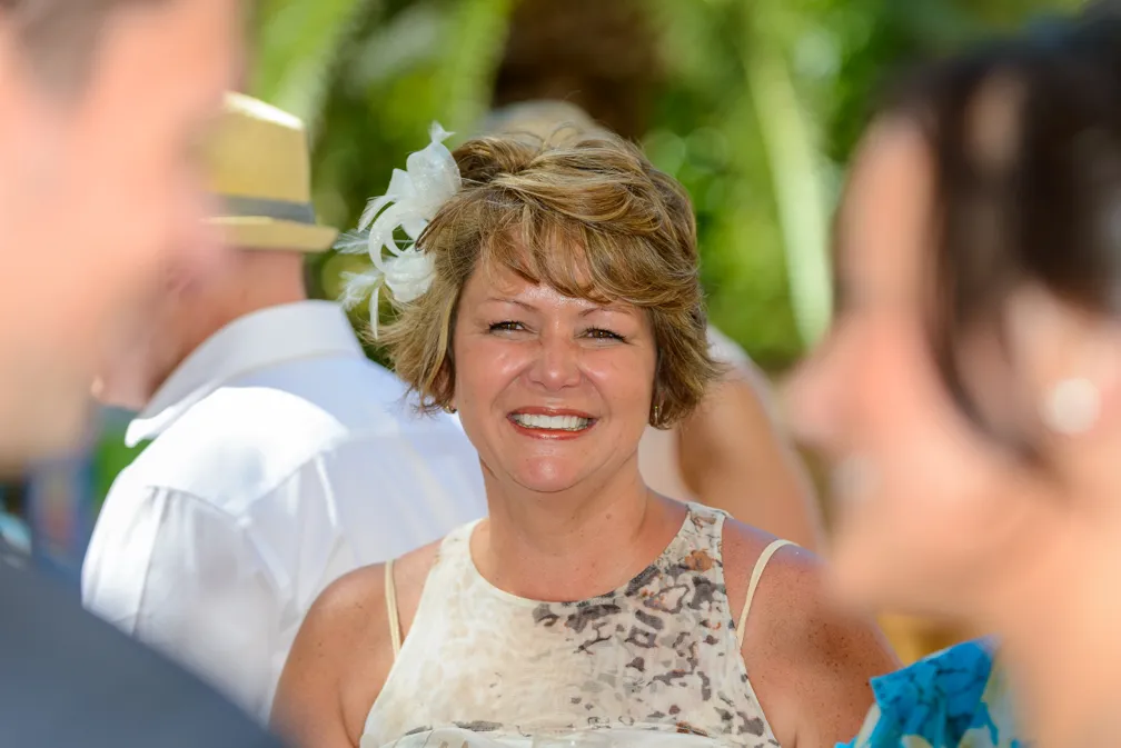 Smiling woman with short blonde hair wearing a sleeveless patterned top and a white decorative hairpiece, with blurred people in the background.