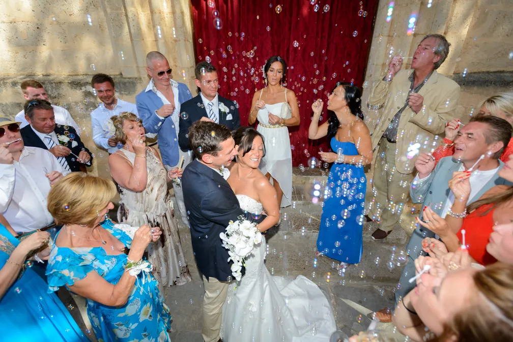 Newlywed couple embracing and smiling surrounded by guests blowing bubbles at an outdoor wedding celebration.