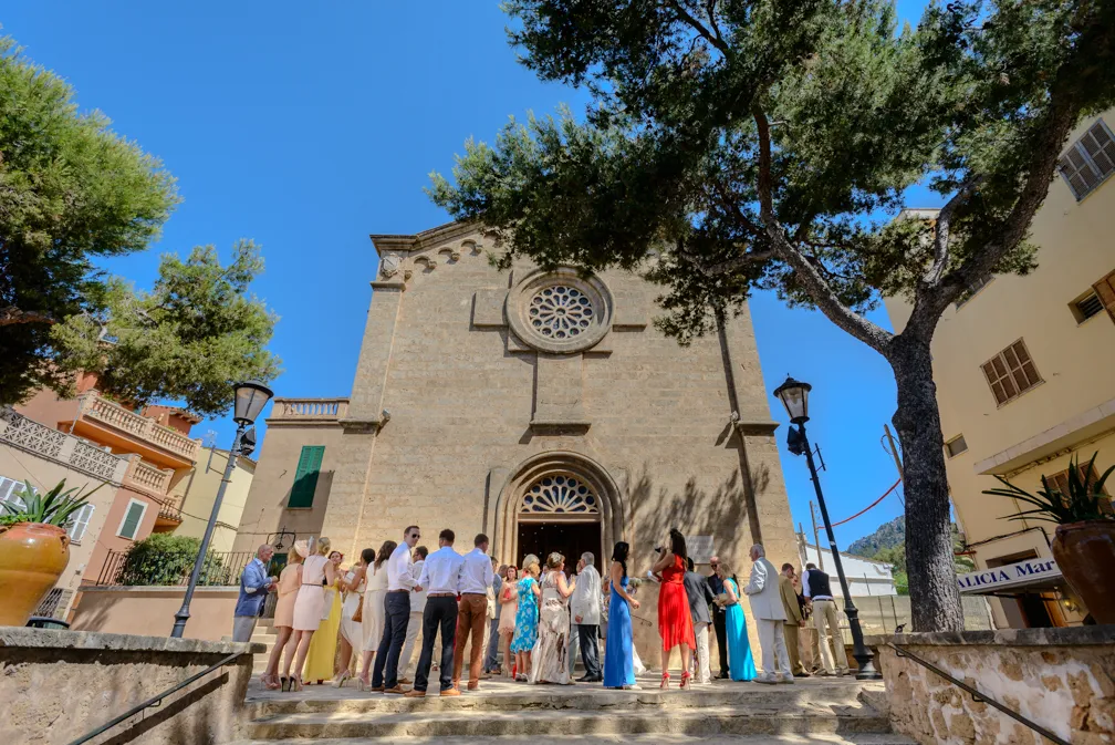 Group of well-dressed people gathered on stone steps outside a historic church with a large rose window under a clear blue sky.