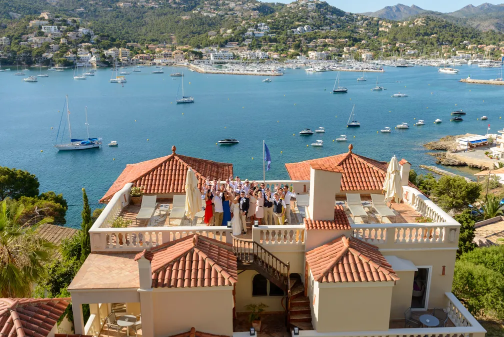 Group of people gathered on a terrace of a building with terracotta roofs overlooking a bay filled with anchored sailboats and yachts.