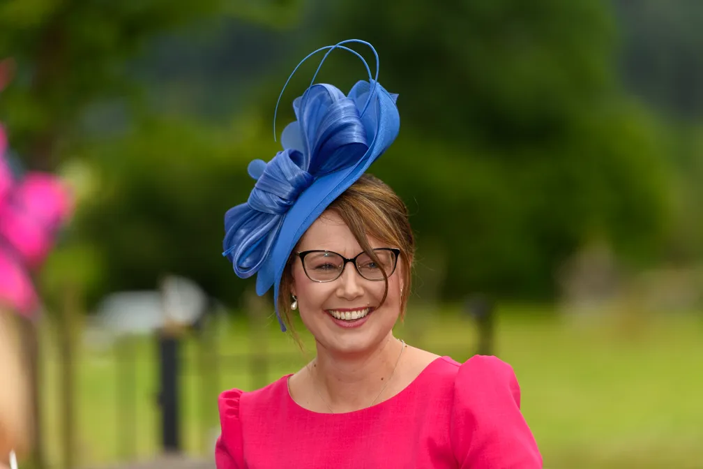 Smiling woman wearing glasses, a bright pink dress, and an elaborate blue fascinator hat outdoors.