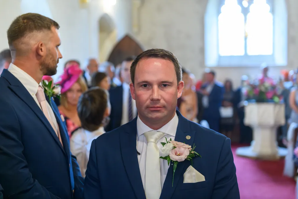 Man in a blue suit with a boutonniere standing in a church during a wedding ceremony.