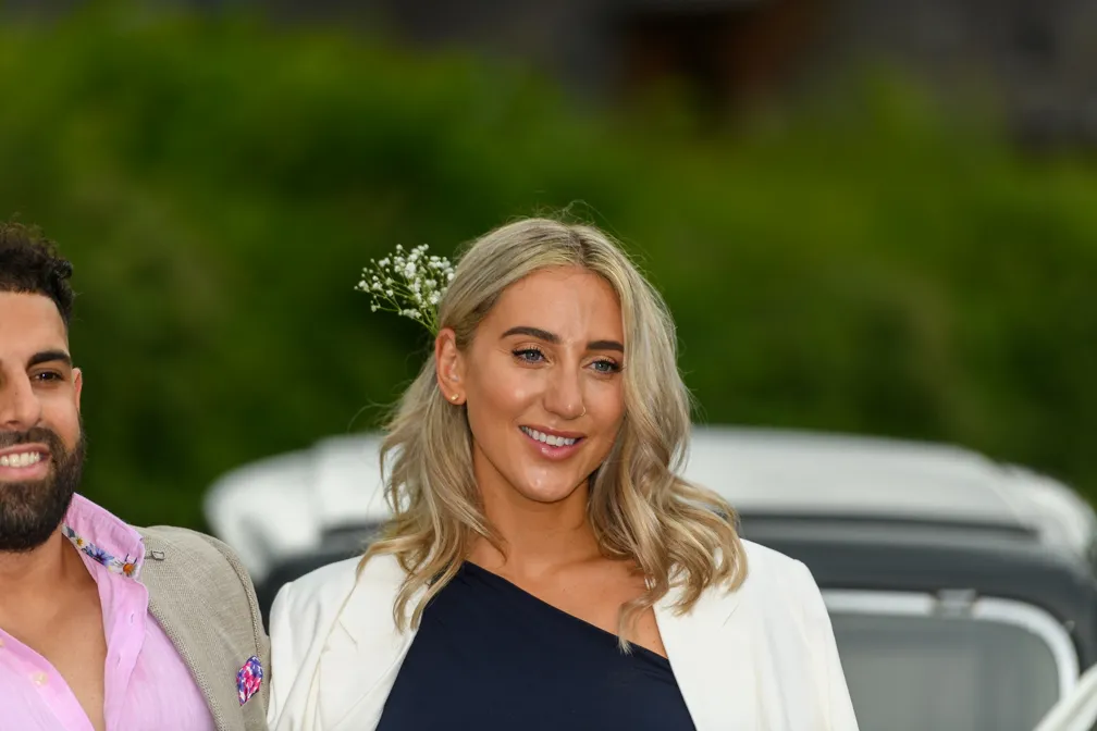 Smiling woman with long blonde hair, wearing a navy blue top and white blazer, with white flowers tucked behind her ear.