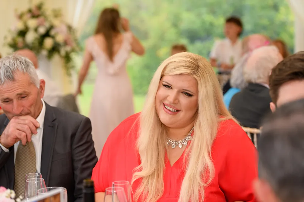Woman with long blonde hair wearing a red blouse and a statement necklace smiling at a social event with seated people in the background.